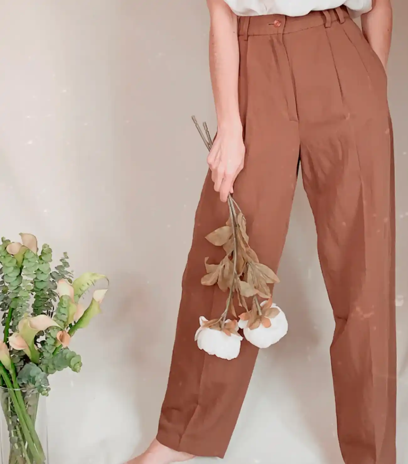 Mujer con pantalón marrón de tiro alto y blusa blanca, sosteniendo ramas decorativas de algodón seco