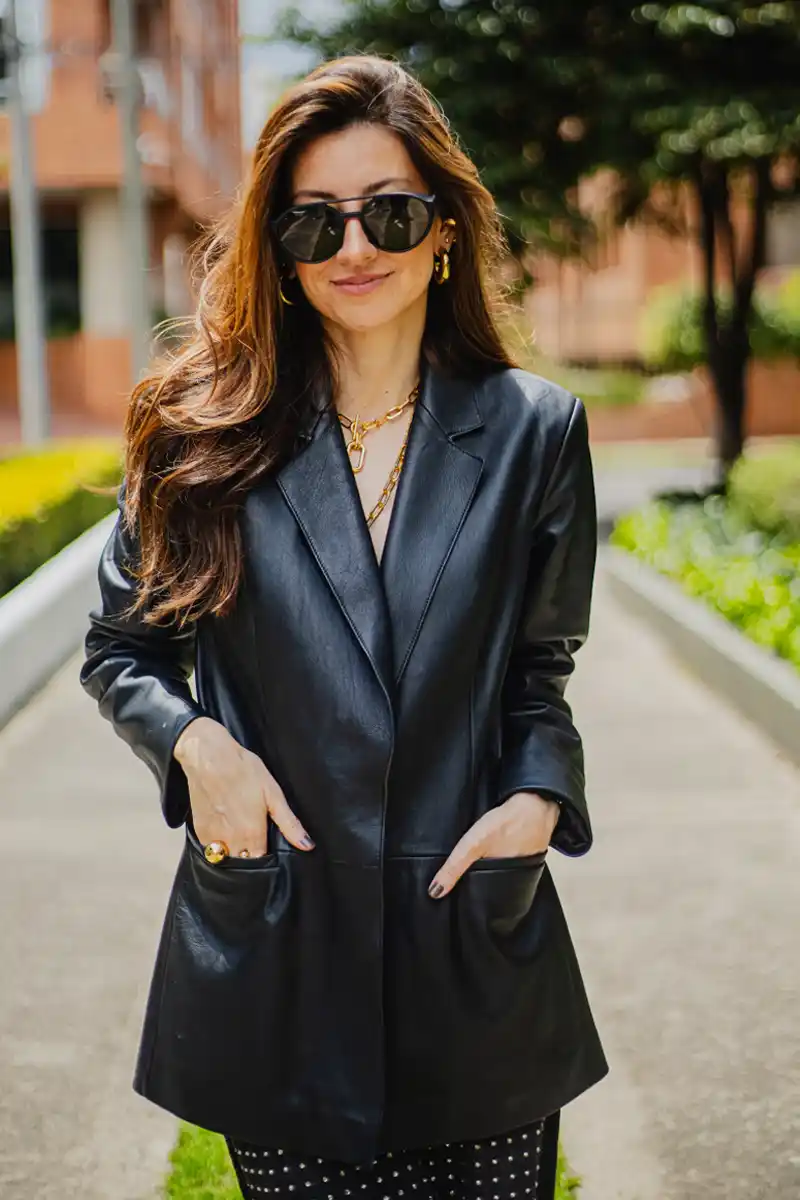 Mujer con blazer negro de cuero posando al aire libre con gafas oscuras y accesorios dorados